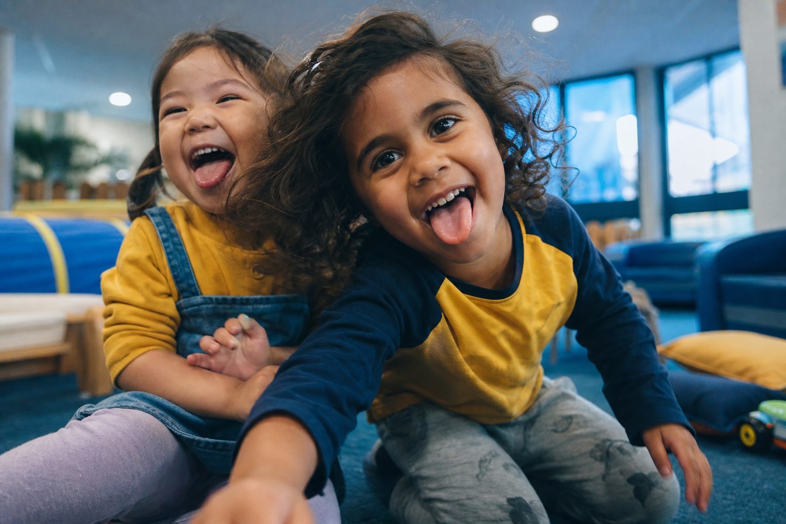 Two children laughing with their tongues out in a bright playroom